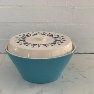 以下が含まれることがあります： 青いセラミック製のサービングボウルと白い蓋。蓋には青と緑の色合いの花柄が描かれています。