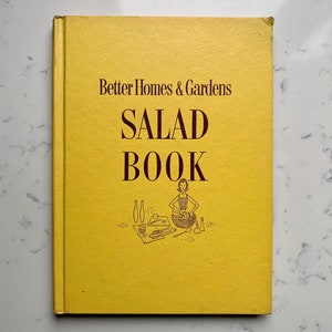 以下が含まれることがあります： 黄色い表紙のヴィンテージ「Better Homes & Gardens SALAD BOOK」。タイトルは大きな濃い茶色の文字で書かれています。女性がサラダを作っているイラストが下にあります。白い大理石の表面に本が置かれています。