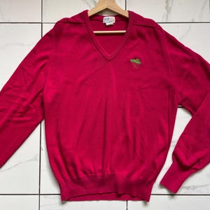 Könnte beinhalten: Ein leuchtend pinkfarbener V-Ausschnitt-Pullover mit langen Ärmeln, der an einem Holzbügel hängt. Der Pullover hat ein kleines grünes, gesticktes Design auf der Brust. Das Markenetikett ist am Ausschnitt sichtbar.