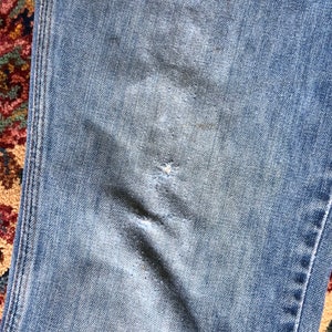 May include: Close-up of light blue denim jeans with visible wear and tear. The jeans show signs of fading and a small tear, suggesting a well-worn appearance. The stitching is visible along the seams.