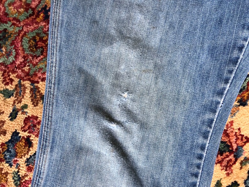 May include: Close-up of light blue denim jeans with visible wear and tear. The jeans show signs of fading and a small tear, suggesting a well-worn appearance. The stitching is visible along the seams.