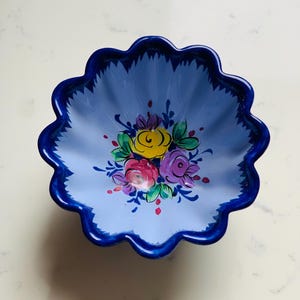 May include: A blue ceramic bowl with a scalloped edge and a floral design. The bowl's interior is light blue, with a dark blue rim. The floral design features yellow, pink, and purple flowers with green leaves.