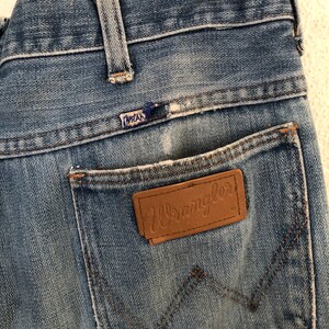 May include: Close-up of vintage blue denim jeans. The jeans feature a brown leather patch with the "Wrangler" logo on the back pocket. The stitching is visible, and the jeans show signs of wear.