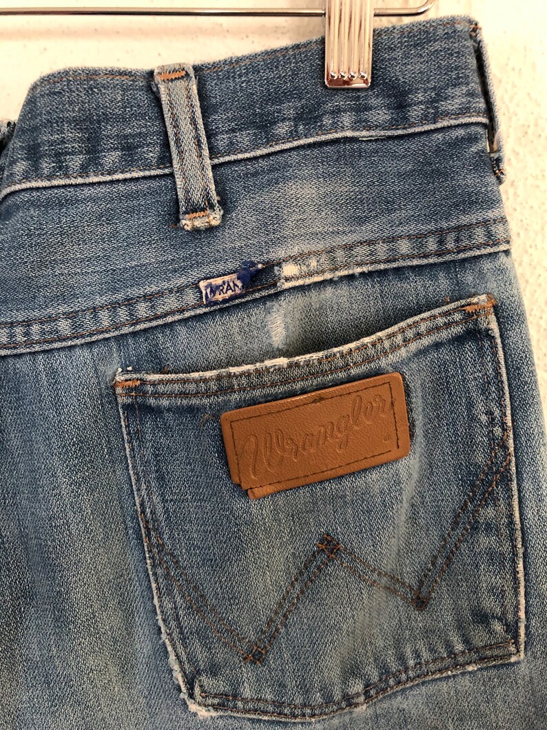 May include: Close-up of vintage blue denim jeans. The jeans feature a brown leather patch with the "Wrangler" logo on the back pocket. The stitching is visible, and the jeans show signs of wear.