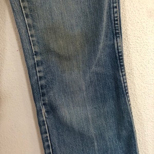 May include: Close-up of a section of blue denim jeans. The fabric shows wear and fading, with a lighter patch on the denim. The stitching is visible along the seam. The jeans are against a white textured background.