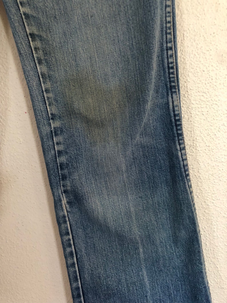 May include: Close-up of a section of blue denim jeans. The fabric shows wear and fading, with a lighter patch on the denim. The stitching is visible along the seam. The jeans are against a white textured background.
