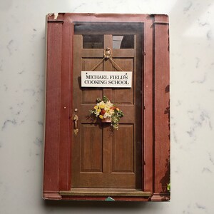 Puede incluir: Portada de libro de cocina con una puerta de madera y un letrero que dice "MICHAEL FIELD'S COOKING SCHOOL". Un arreglo floral cuelga debajo del letrero. La puerta está enmarcada por una estructura marrón rojiza, que sugiere una entrada a la casa.