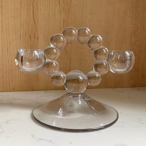 May include: Clear glass candle holder with two cup-shaped candle holders and a circular design of glass spheres. The candle holder sits on a wide, circular base. The background is a light brown wooden surface.