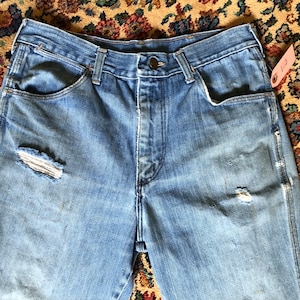 May include: Light blue denim jeans with distressed details, including a ripped knee. The jeans have a button closure and classic five-pocket design. A pink tag is attached to the jeans. The jeans are vintage.