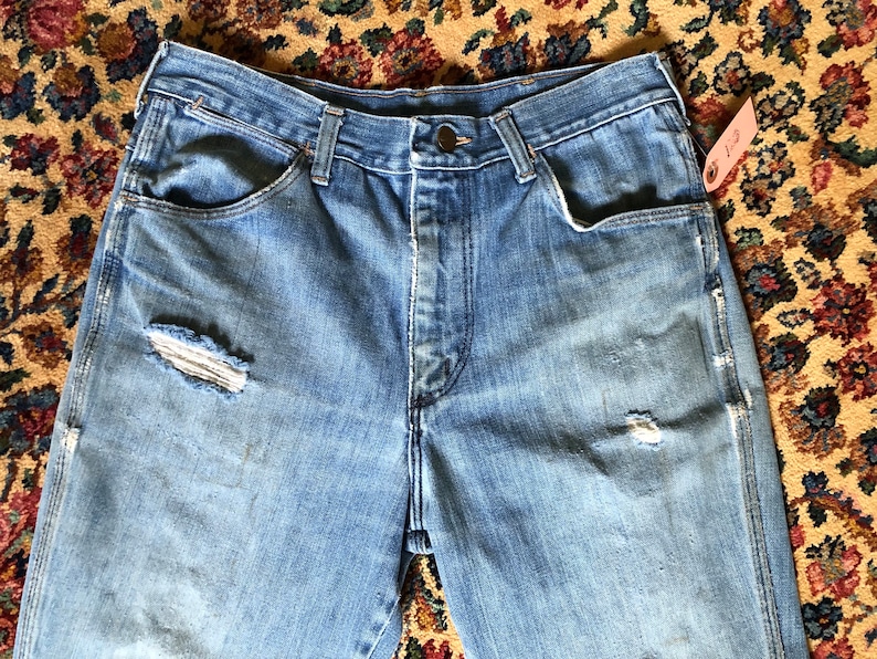 May include: Light blue denim jeans with distressed details, including a ripped knee. The jeans have a button closure and classic five-pocket design. A pink tag is attached to the jeans. The jeans are vintage.