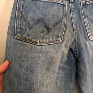 May include: Close-up of light blue denim jeans with a Wrangler logo on the back pocket. The jeans show signs of wear, with some staining and distressing. The denim has a faded appearance.