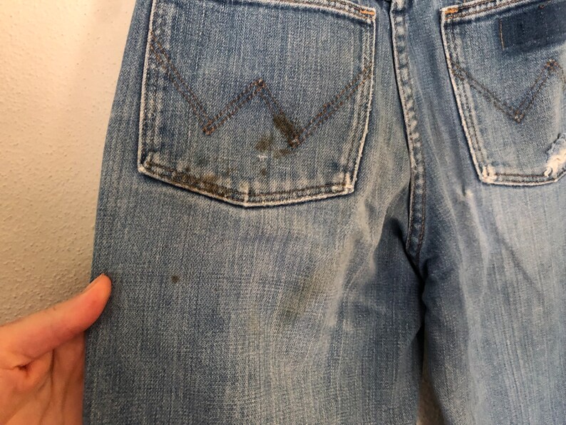 May include: Close-up of light blue denim jeans with a Wrangler logo on the back pocket. The jeans show signs of wear, with some staining and distressing. The denim has a faded appearance.