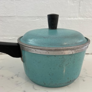 Puede incluir: Una cacerola turquesa vintage con tapa y asa negra. La olla tiene un borde plateado y un pomo negro en la tapa. La cacerola es de metal y está sobre una superficie blanca.