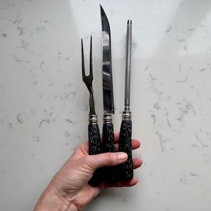 May include: A carving set with a fork, knife, and sharpening steel. The knife has a curved blade. The handles are dark brown with a textured pattern, and the metal parts are silver. The set is designed for carving meat.