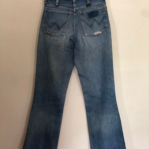 May include: Vintage blue denim jeans with a classic bootcut leg. The jeans feature the Wrangler logo on the back pockets and a worn, distressed look. The jeans are hanging on a wooden hanger.