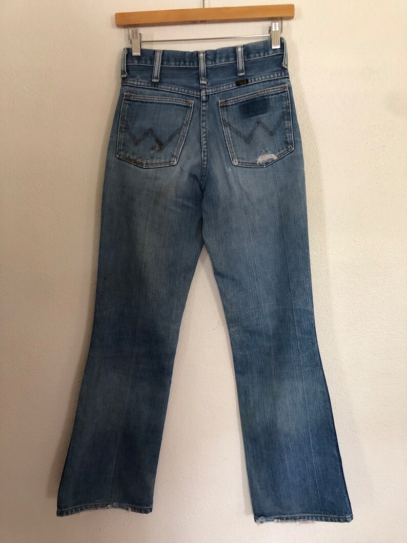 May include: Vintage blue denim jeans with a classic bootcut leg. The jeans feature the Wrangler logo on the back pockets and a worn, distressed look. The jeans are hanging on a wooden hanger.