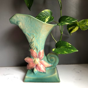May include: A turquoise ceramic vase with a flared top and a square base. The vase features a sculpted pink and yellow flower and leaf design. The vase is approximately 10 inches tall.