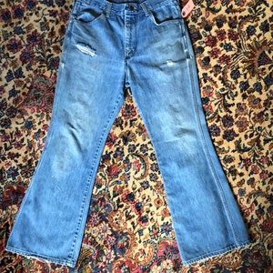 May include: Light blue denim jeans with a flared leg design. The jeans have a distressed look with a ripped knee and faded areas. A small tag is attached to the side. The jeans are laid flat on a patterned rug.