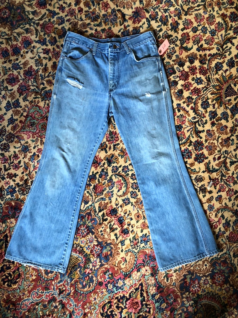 May include: Light blue denim jeans with a flared leg design. The jeans have a distressed look with a ripped knee and faded areas. A small tag is attached to the side. The jeans are laid flat on a patterned rug.