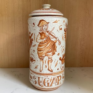 May include: A ceramic sugar jar with a lid, featuring a tan and white crackle glaze. The jar is decorated with a violinist figure and the word "SUGAR" in a decorative font. The jar is cylindrical and has a matching lid.