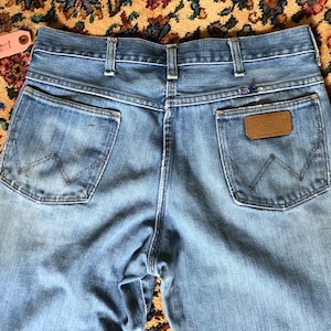 May include: Light blue denim jeans with a classic five-pocket design. The back pockets feature the iconic Wrangler stitching. A brown leather patch with the Wrangler logo is visible. The jeans are vintage and show signs of wear.