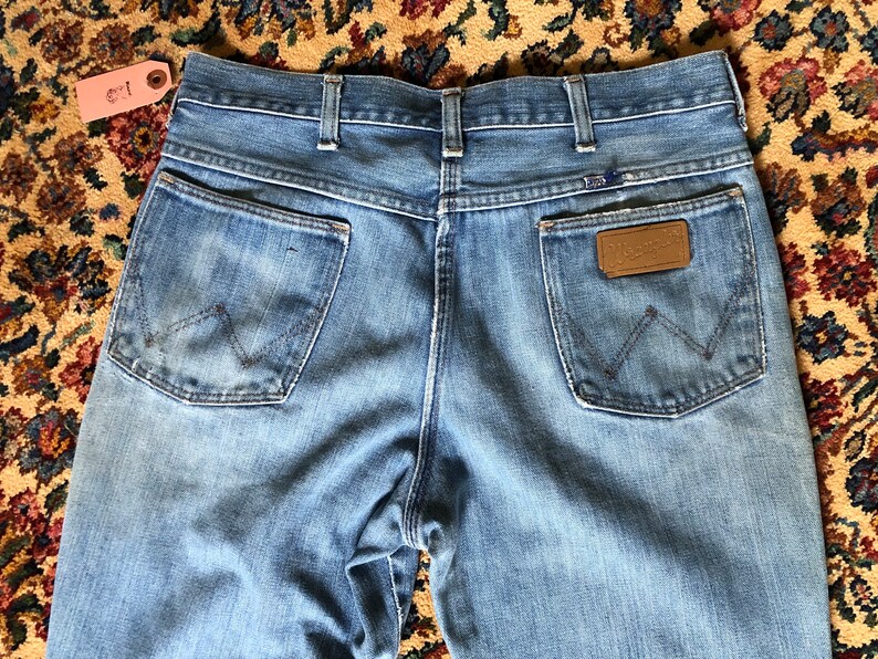 May include: Light blue denim jeans with a classic five-pocket design. The back pockets feature the iconic Wrangler stitching. A brown leather patch with the Wrangler logo is visible. The jeans are vintage and show signs of wear.