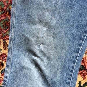 May include: Close-up of light blue denim jeans with distressed details. The jeans show wear and tear with a visible rip and faded patches, suggesting a vintage or worn aesthetic. The fabric has a classic denim texture.