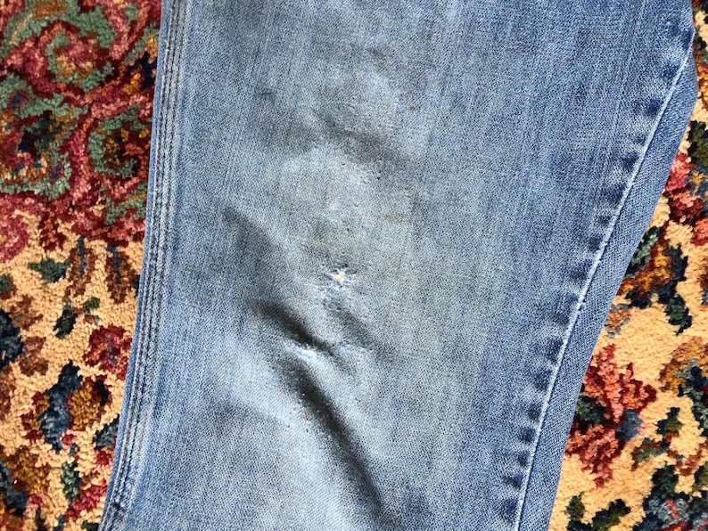 May include: Close-up of light blue denim jeans with distressed details. The jeans show wear and tear with a visible rip and faded patches, suggesting a vintage or worn aesthetic. The fabric has a classic denim texture.