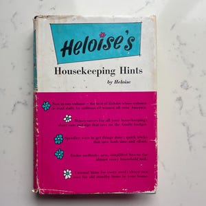 May include: A vintage book titled "Heloise's Housekeeping Hints" with a white and turquoise top section and a pink bottom section. The book cover features the title in black and the author's name. Decorative floral elements are scattered on the pink section.