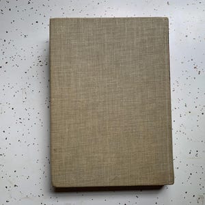 May include: A rectangular book with a textured, tan fabric cover. The book is lying flat on a white surface with small black speckles. The cover has a simple, classic design.