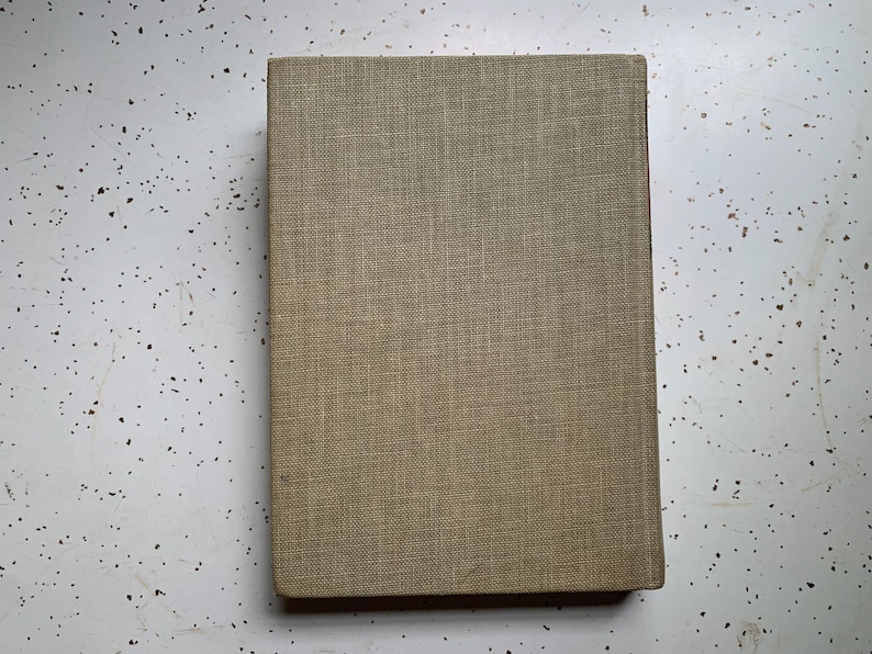 May include: A rectangular book with a textured, tan fabric cover. The book is lying flat on a white surface with small black speckles. The cover has a simple, classic design.