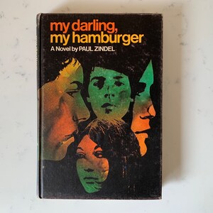 May include: Book cover with the title "my darling, my hamburger" by Paul Zindel. The cover features a black background with four faces in profile, two male and two female, in shades of orange, green, and yellow.