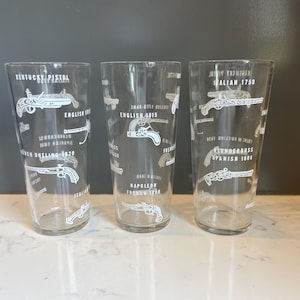 May include: Three clear drinking glasses with white illustrations of antique pistols and text. The glasses feature different pistol designs and text such as "Kentucky Pistol" and "Italian 1750". The glasses are on a white surface.