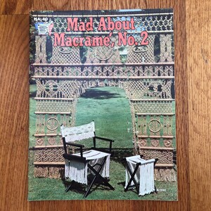 May include: Cover of "Mad About Macramé, No. 2" magazine. The cover features a macramé archway and a chair and stool set. The chair and stool have black frames and white macramé seats and fringe. The archway is made of beige macramé.