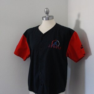 May include: Black baseball jersey with red sleeves. Features a button-up front and a logo on the chest with the word "Jordan" below it. The Nike logo is visible on the right sleeve.