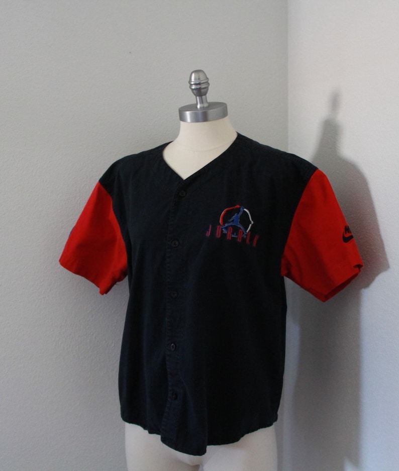 May include: Black baseball jersey with red sleeves. Features a button-up front and a logo on the chest with the word "Jordan" below it. The Nike logo is visible on the right sleeve.