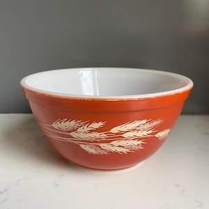 Puede incluir: Un cuenco mezclador Pyrex naranja vintage con interior blanco. El exterior presenta un diseño de trigo en color crema. El cuenco es redondo y tiene un diseño clásico, perfecto para la cocina.