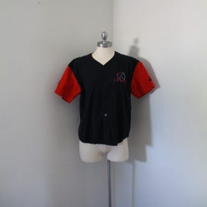 May include: Black baseball jersey with red sleeves. The jersey has a V-neck and button-up front. A small logo is embroidered on the chest. The sleeves have a Nike logo. The jersey is displayed on a mannequin.