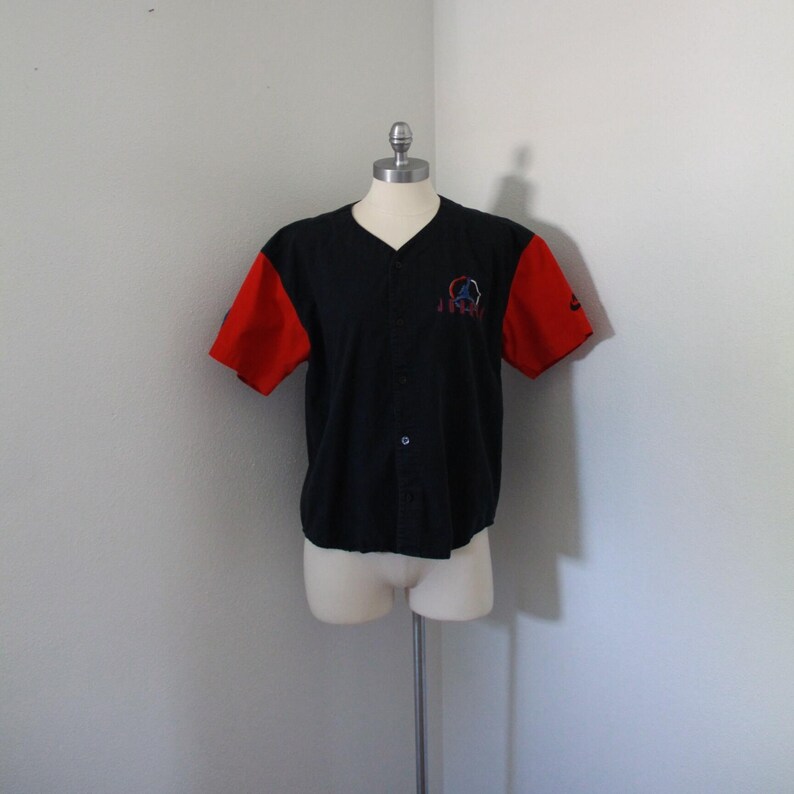 May include: Black baseball jersey with red sleeves. The jersey has a V-neck and button-up front. A small logo is embroidered on the chest. The sleeves have a Nike logo. The jersey is displayed on a mannequin.