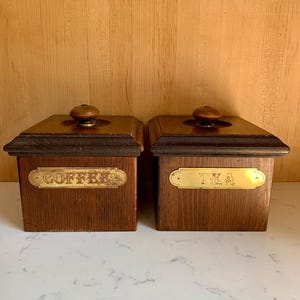 May include: Two wooden storage boxes with brass labels. One reads "COFFEE" and the other "TEA". Each box has a dark wooden lid with a round, wooden knob. The boxes are brown and sit on a white surface.