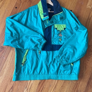 May include: Teal and navy blue anorak jacket with neon green accents. The jacket features a half-zip closure, a collared neckline, and a front pocket. The sleeves have elastic cuffs. The jacket is hanging on a wooden hanger.