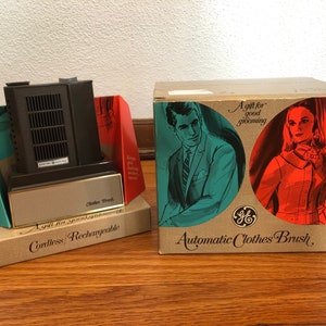 May include: Vintage General Electric Automatic Clothes Brush in original packaging. The box features illustrations of a man and woman. The brush is black and gold, with the words "Cordless/Rechargeable" on the box.