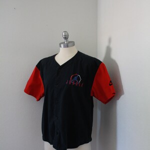 May include: Black baseball jersey with red sleeves. The jersey features a v-neck, button-up front, and an embroidered logo on the chest. The Nike logo is visible on the right sleeve.