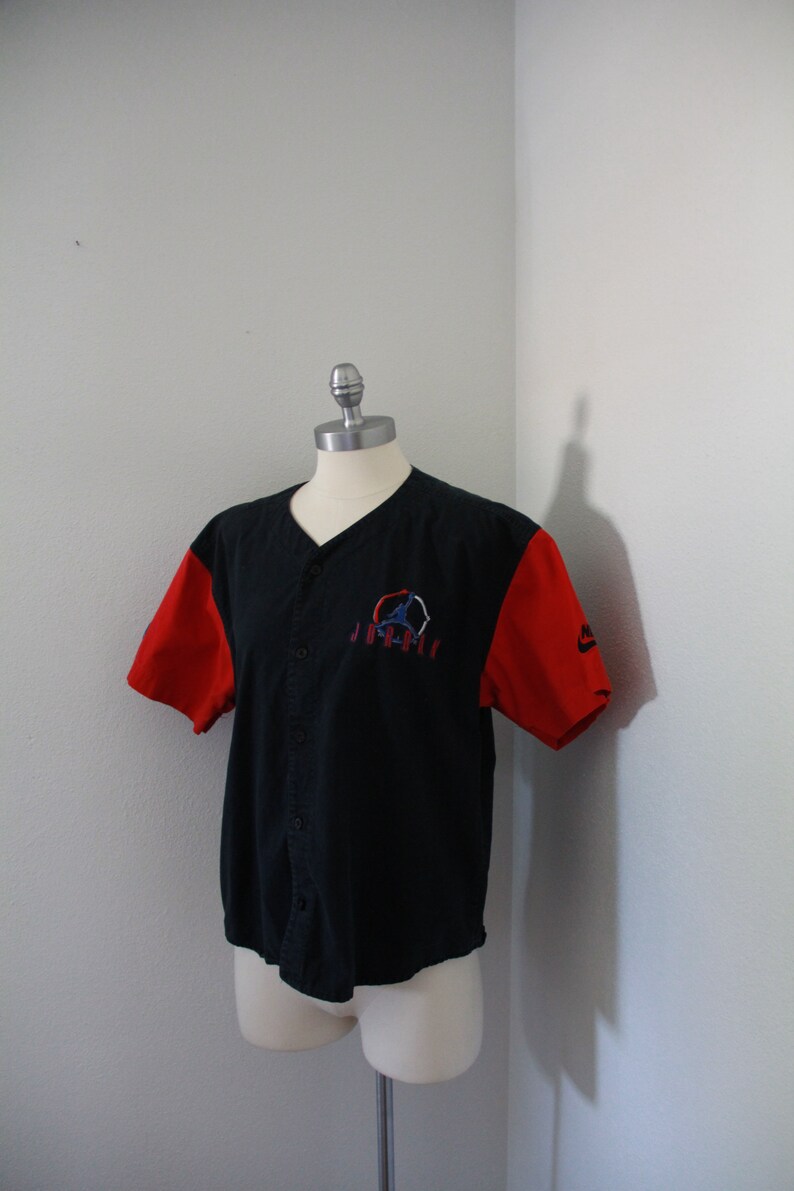 May include: Black baseball jersey with red sleeves. The jersey features a v-neck, button-up front, and an embroidered logo on the chest. The Nike logo is visible on the right sleeve.