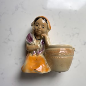Może przedstawiać: Figurka ceramiczna w stylu vintage przedstawiająca kobietę w kolorowej sukience, opartą o beżowy garnek. Kobieta ma na sobie pomarańczową chustę, fioletową szal i pomarańczową spódnicę. Garnek ma ozdobny pas pośrodku.