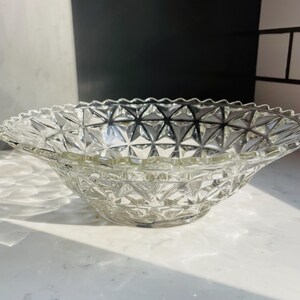 Op de afbeelding: Transparante glazen kom met een ruitpatroon en een geschulpte rand. De kom heeft een brede, ondiepe vorm, geschikt om te serveren of tentoon te stellen. Het transparante glas laat licht door, waardoor het geometrische ontwerp wordt benadrukt.