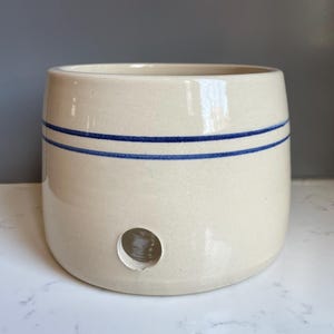 Puede incluir: Olla de cerámica color crema con dos rayas azules horizontales y un orificio de drenaje. El recipiente cilíndrico tiene una superficie ligeramente texturizada. Esta olla de estilo vintage es adecuada para la cocina o la decoración del hogar.