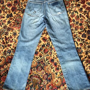 May include: Light blue denim jeans with a faded wash and distressed details. The jeans feature classic five-pocket styling, a zip fly, and the Wrangler logo on the back pockets. The hem is frayed.