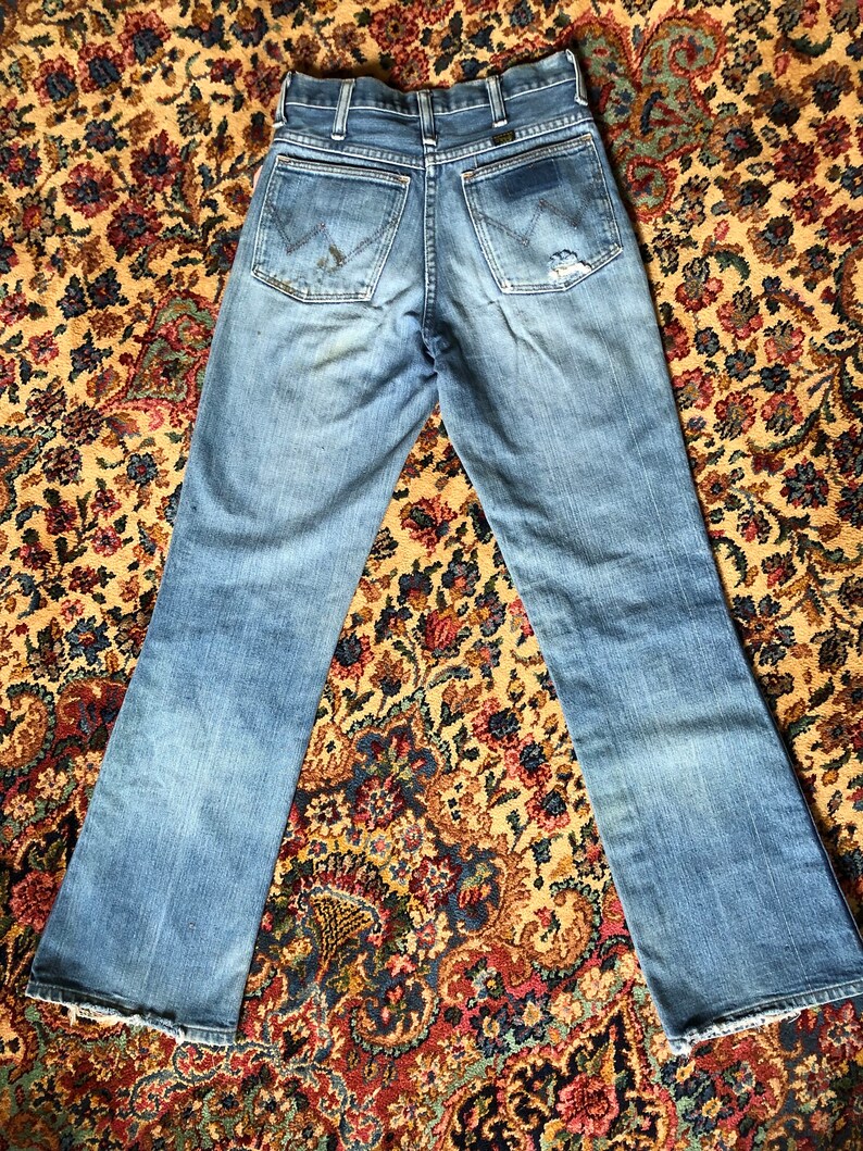 May include: Light blue denim jeans with a faded wash and distressed details. The jeans feature classic five-pocket styling, a zip fly, and the Wrangler logo on the back pockets. The hem is frayed.