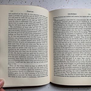 May include: Open book with text on the pages. The left page has the title "Emerson" at the top, and the right page has the title "Self-Reliance". The book is open to pages 122 and 123.